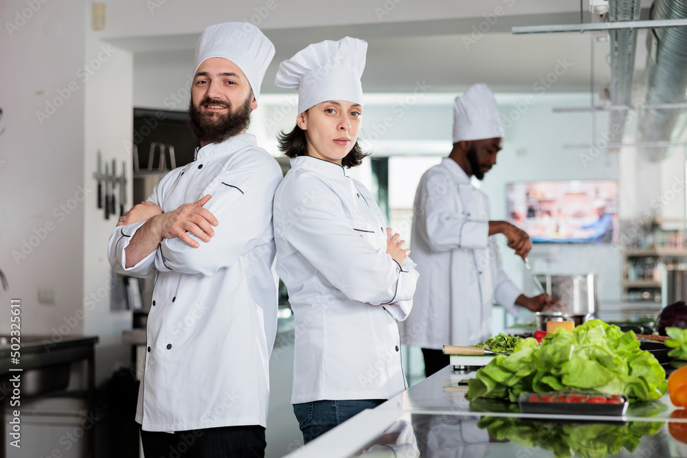 Back to back gastronomy experts standing in restaurant professional kitchen while posing for camera. Chefs wearing cooking uniformswhile standing in gourmet cuisine with arms crossed.