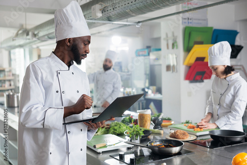 Gastronomy expert wearing professional cooking uniform while searching meal ideas. Gourmet cuisine head chef with laptop looking for gastronomic dish recipes and garnish ideas in restaurant kitchen.