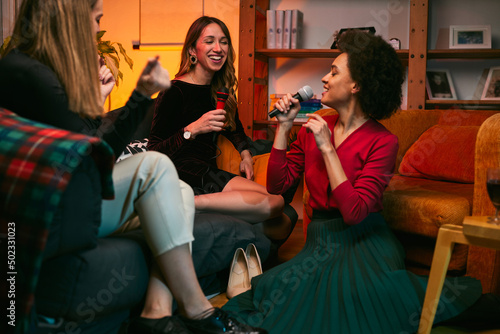 A multicultural group of female friends sings karaoke while drinking wine in the living room. © chika_milan