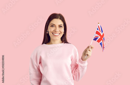 Portrait of happy smiling young woman with flag of UK. Pretty British girl with cheerful face expression standing isolated on pink background and holding Union Jack. Learning English language concept photo