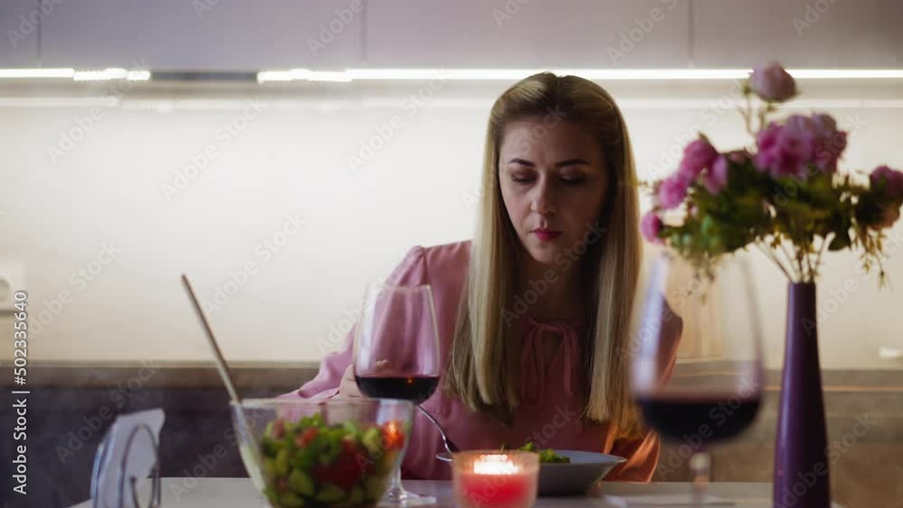 Pretty blonde woman in stylish dress with tasty red wine glass eats fresh juicy salad sitting at table at festive dinner in kitchen slow motion Pretty blonde woman in stylish dress with tasty red wine glass eats fresh juicy salad sitting at table at festive dinner in kitchen slow motion