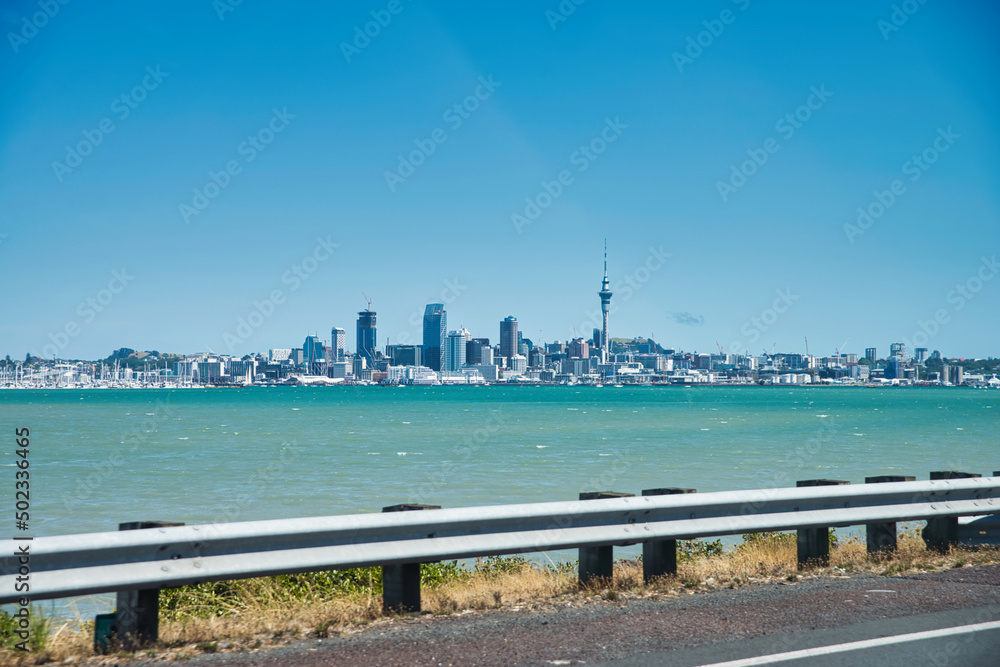 Auckland skyline on a sunny day, seen from a highway across the bay with parts of the street and the foreground and lots of blue turquoise water in between. 