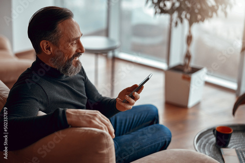 Happy man sitting in armchair and looking at smartphone near big window, copy space