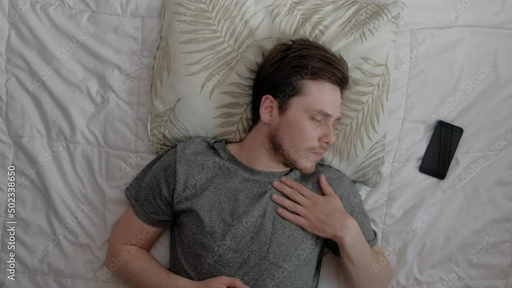Top view of handsome sleepy man is sleeping in the bed, he wake up for looking how time on smartphone, and decides to get some more sleep. Top view of handsome sleepy man is sleeping in the bed, he wake up for looking how time on smartphone, and decides to get some more sleep.