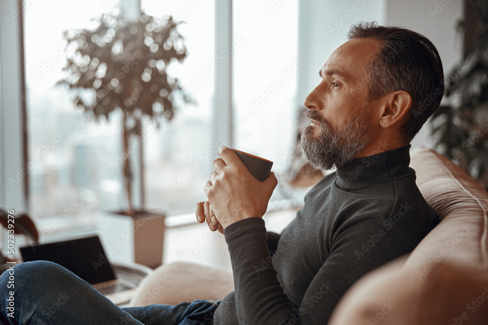 Side view of adult man resting and holding cup of coffee while looking out the window Side view of adult man resting and holding cup of coffee while looking out the window