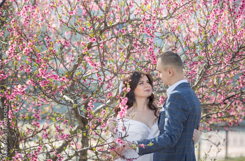 a young girl in a white dress and a man in a suit stand by a flowering tree with pink flowers and look at each other..