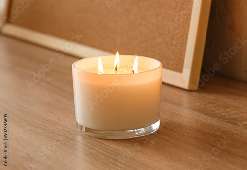 A large scented candle with three burning wicks and melting wax in a glass, standing on a wooden table against the background of a board with cork notes. Side view. Place to copy
