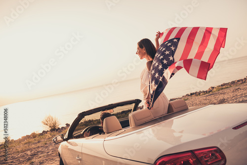 Photo of funny carefree married couple dressed white clothes driving car holding american flag outdoors country side road