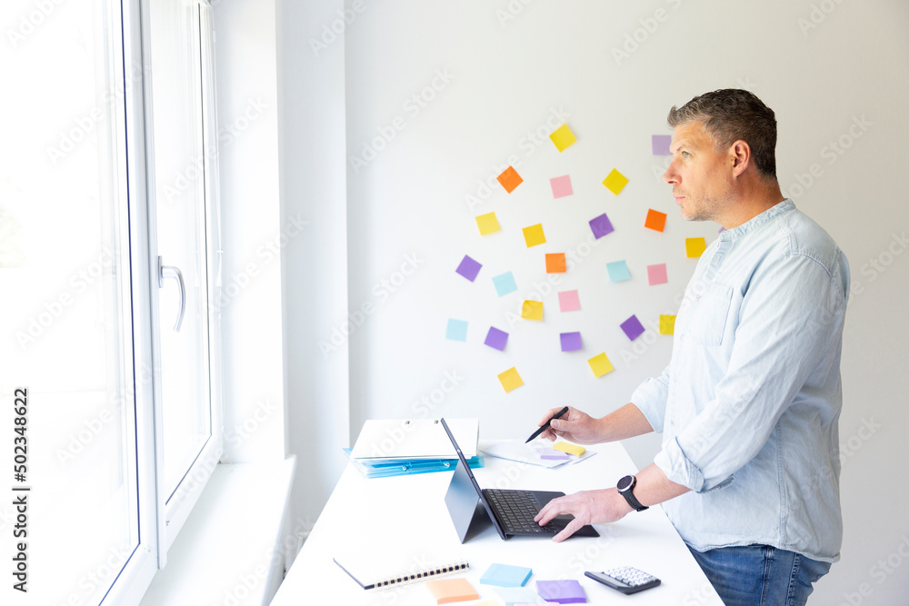 vMan with blue shirt stands at white height adjustable table and works on notebook behind him on the wall stick colorful notes vMan with blue shirt stands at white height adjustable table and works on notebook behind him on the wall stick colorful notes
