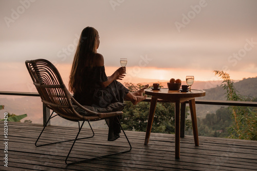 Young woman with a glass of white wine on the terrace overlooking the sunset in the mountains