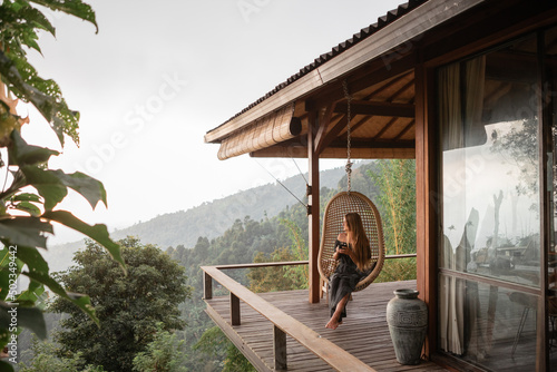 Tourist woman swing on wicker rattan hang chair in the jungle, nature mountains view, hold in hands cup of tea/coffee © Yevhenii