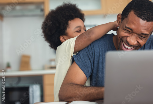 Cute little African American boy hugging and playing with dad while his dad working from home with laptop computer for remoter job in modern kitchen. Concepts of remote work, children, parenthood