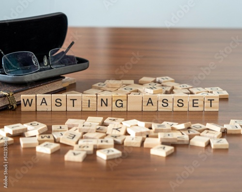 wasting asset word or concept represented by wooden letter tiles on a wooden table with glasses and a book