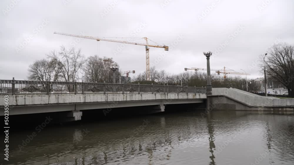 Concrete wet bridge across city river during rainy season with high water levels. Cloudy day with trees without leaves and idle tower cranes. Concrete wet bridge across city river during rainy season with high water levels. Cloudy day with trees without leaves and idle tower cranes.