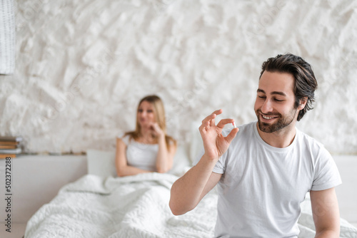 Young man holding pill, solving sexual dysfunction problems, happy woman sitting in bed and waiting for her husband photo