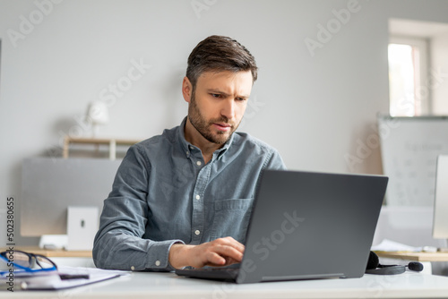 Mature male manager working on laptop at modern office, sitting at table, typing on computer keyboard