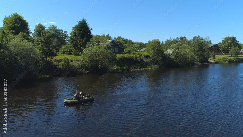 A men in a rubber boat rowing along the river bank in the village. Shot. The concept of tourism, vacation, hobbies and outdoor activities, two men sailing in a boat near wooden house and green trees
