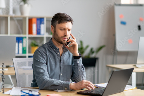 Focused mature businessman having phone conversation with client, sitting at office, typing on laptop keyboard