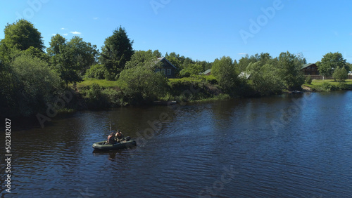 A men in a rubber boat rowing along the river bank in the village. Shot. The concept of tourism, vacation, hobbies and outdoor activities, two men sailing in a boat near wooden house and green trees