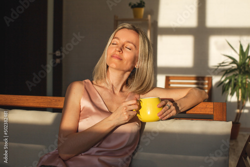 Obraz na plátně Happy dreamy middle aged woman sitting on sofa in living room with cup of tea or