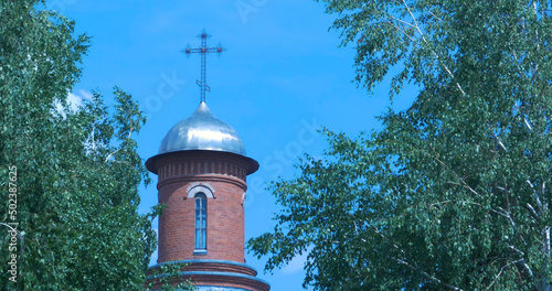 Orthodox temple orange brick with sunny ray, flowering green trees in foreground. Blue sky white clouds. Pray, holy light new christ church. Summer time, sunny day photo