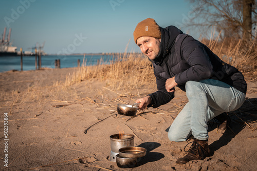 Young white man an his knees with warm clothes on is cooking with outdoor cooker on Harriersand beach at the riverbank of Weser during golden hour. Harbor and ships blurred in the background. photo