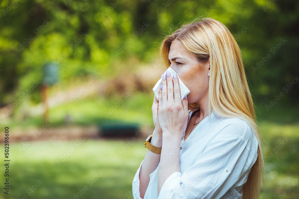 Young pretty woman blowing nose in front of blooming tree. Spring allergy concept. Attractive young adult woman coughing and sneezing outdoors. Sick people allergy or virus influenca concept. Young pretty woman blowing nose in front of blooming tree. Spring allergy concept. Attractive young adult woman coughing and sneezing outdoors. Sick people allergy or virus influenca concept.