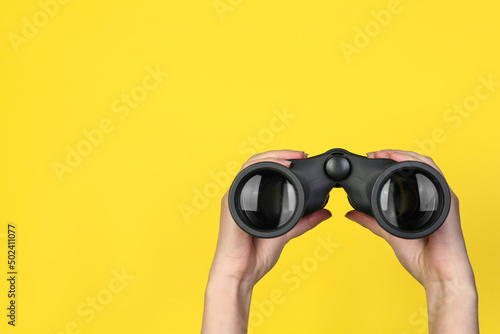 Woman holding modern binoculars on yellow background, closeup. Space for text photo