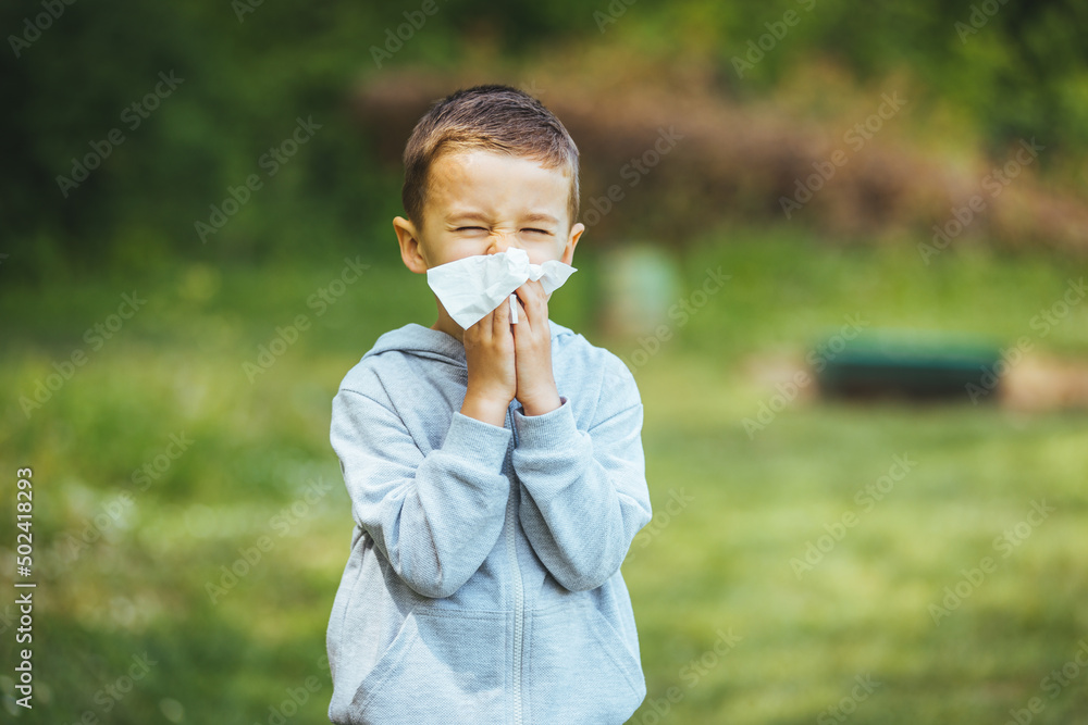 Boy has allergies from flower pollen. Allergy concept. Little boy is blowing her nose near blooming tree. Little boy using tissue from allergy infection in the park