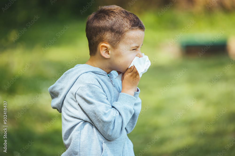 Little blond boy sneezing due to allergy related problems, on a sunny day outdoors. He is holding a handkerchief in his hands, looking away. Copy space available. Little blond boy sneezing due to allergy related problems, on a sunny day outdoors. He is holding a handkerchief in his hands, looking away. Copy space available.