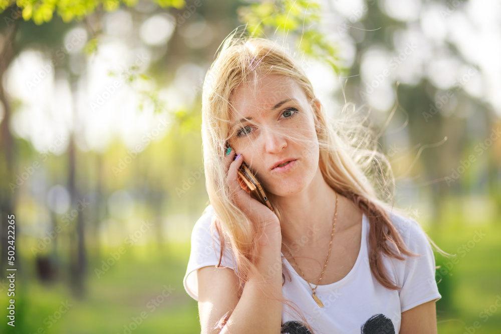 Blonde girl talking on the phone in the park in spring. Portrait of a beautiful fashionista mom with long hair talking to a friend on a smartphone.
