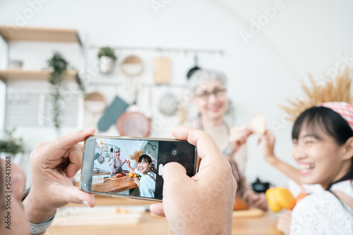 Elderly man having fun taking a family photo with his smartphone.Asian parents and daughter eating together on the dining table in a cozy kitchen.Happy time using technology.Thai,Celebration,Eve.