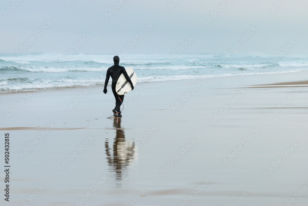 Surfer in full wetsuit walks along the beach at sunrise. Landes Surfer in full wetsuit walks along the beach at sunrise. Landes