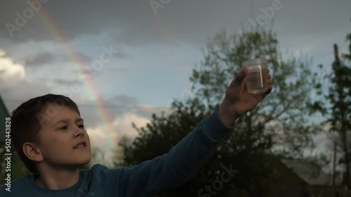 a boy collects rainwater drops in a glass while standing against a rainbow background.on a country plot