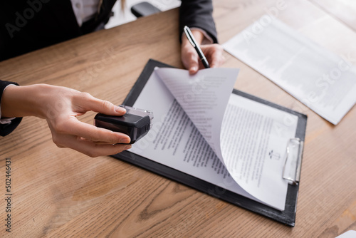 cropped view of lawyer holding stamper and pen near contract on clipboard.