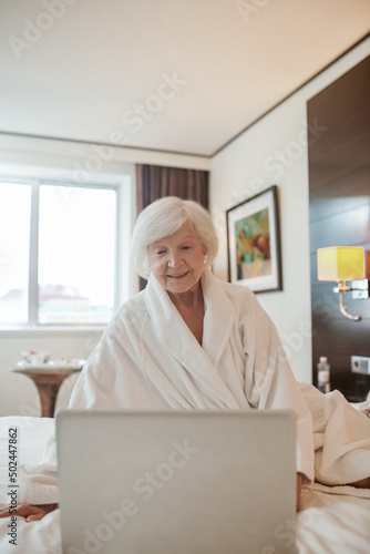 An old woman with a laptop sitting on a bed in a hotel room