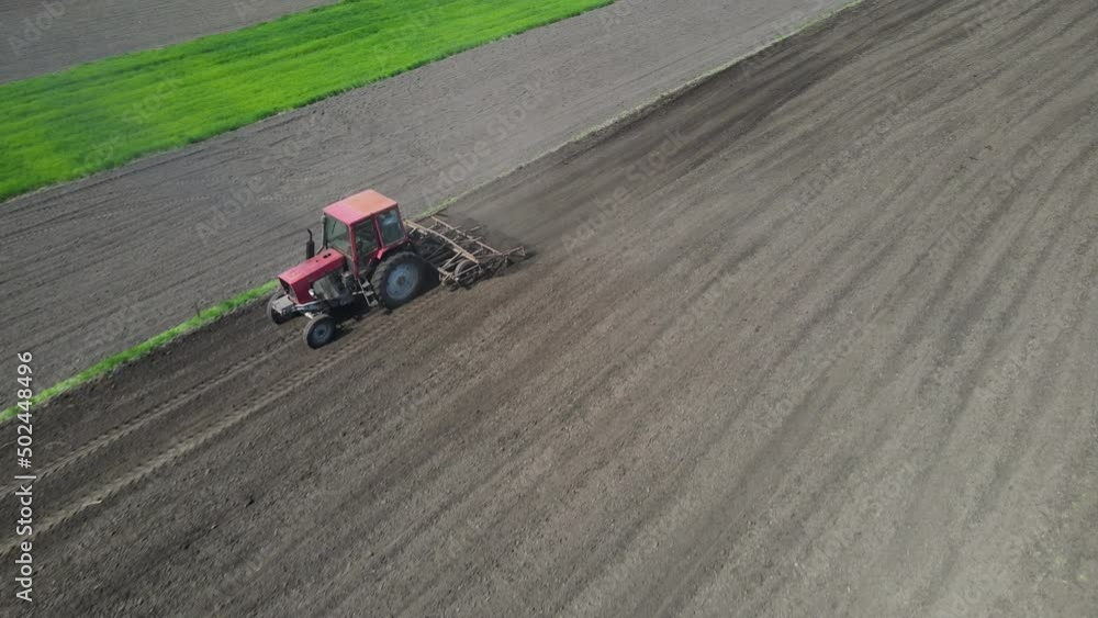 Agricultural red small tractor in the field plowing. System plowing ground on cultivated farm field, pillar of dust trails behind, preparing soil for planting new crop, agriculture concept, top view Agricultural red small tractor in the field plowing. System plowing ground on cultivated farm field, pillar of dust trails behind, preparing soil for planting new crop, agriculture concept, top view