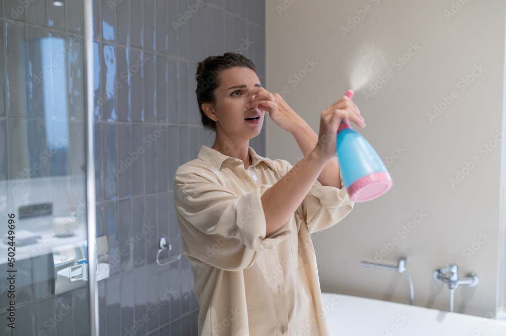 A young woman closing her nose from unpleasant smell and spraying air freshener - obrazy, fototapety, plakaty 
