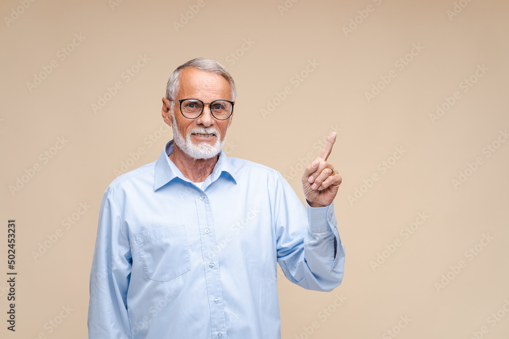 Fototapeta premium Elderly businessman with happy face pointing up with finger at copy space. Indoor studio shot isolated on beige background