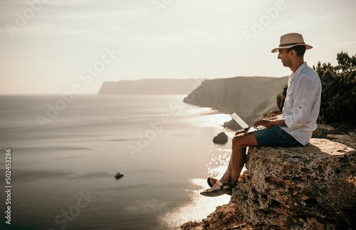 Digital nomad, man in the hat, a businessman with a laptop sits on the rocks by the sea during sunset, makes a business transaction online from a distance. Remote work on vacation.