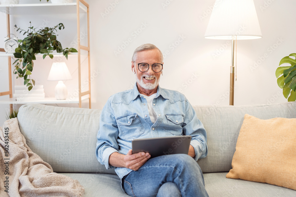 Cheerful senior man wearing casual wear sitting on the sofa and using laptop at modern home office, glad male entrepreneur typing on keyboard
