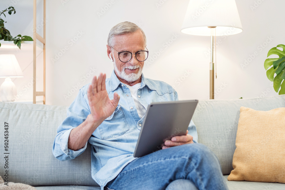 Smiling cheerful happy senior man waving and gesturing while having online video call on hand held tablet portable device talking to and connecting with family or friends Smiling cheerful happy senior man waving and gesturing while having online video call on hand held tablet portable device talking to and connecting with family or friends