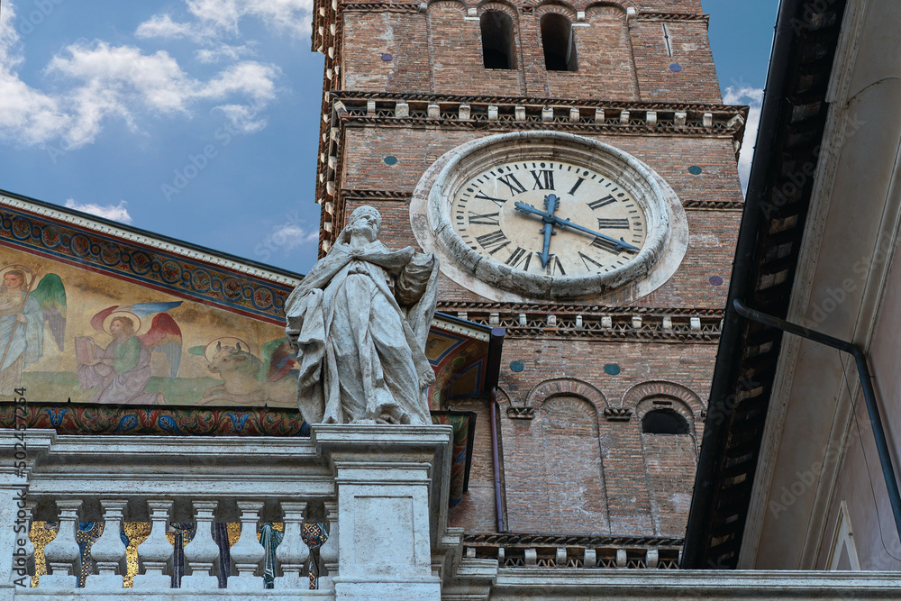a fragment of the facade of a Roman cathedral with a beautiful tower clock and a statue of a saint against the blue sky