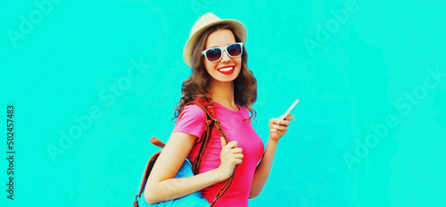 Portrait of happy smiling young woman tourist or student with smartphone wearing summer straw hat, backpack on blue background, blank copy space for advertising text