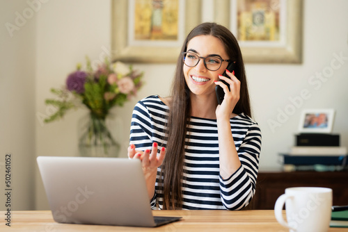 Attractive young woman using laptop and mobile phone while sitting at desk at home photo