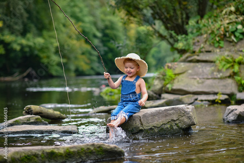 Fototapeta Naklejka Na Ścianę i Meble -  Funny happy little kid fishing at the lake. A fisher boy sits in the lake with a fishing rod and catches fish