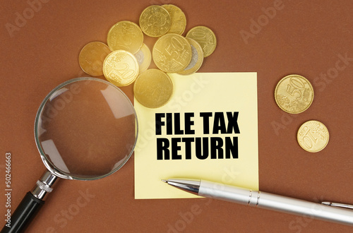On a brown surface are coins, a pen, a magnifying glass and stickers with the inscription - File Tax Return