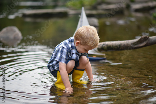 Fototapeta Naklejka Na Ścianę i Meble -  Happy little boy in yellow rain boots playing with ship boat in lake on spring or autumn day. Active leisure for children. Funny kid having fun outdoors wearing colorful clothes.