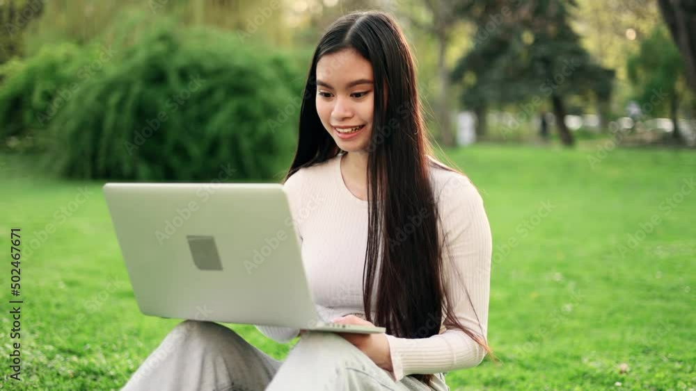 Dreamy attractive asian woman sitting on a grass in the park and talking on laptop by video chat. Student learning online. Dreamy attractive asian woman sitting on a grass in the park and talking on laptop by video chat. Student learning online.