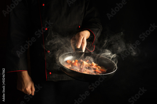 Fototapeta Naklejka Na Ścianę i Meble -  The cook prepares food in a frying pan with steam on a black background. The concept of restaurant and hotel service. European cuisine.